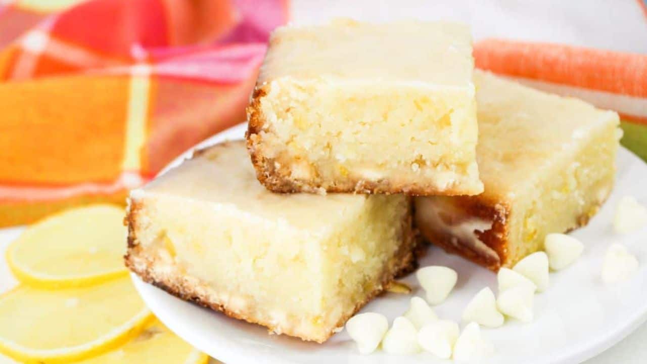 Three lemon bars stacked on a white plate, surrounded by white chocolate chips and lemon slices, with a colorful cloth in the background.