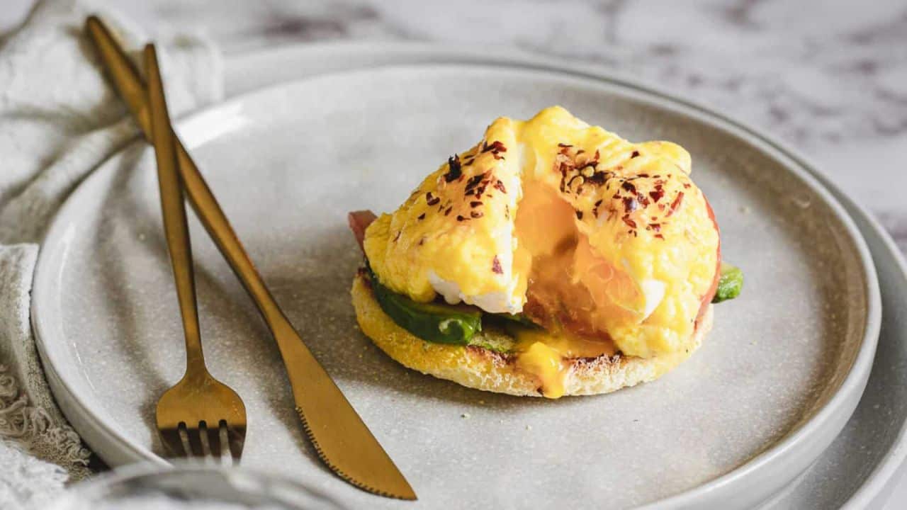 A plate with eggs benedict featuring a poached egg, hollandaise sauce, and avocado slices on an english muffin, garnished with spices, accompanied by gold cutlery. Perfect for Mother's Day recipes.