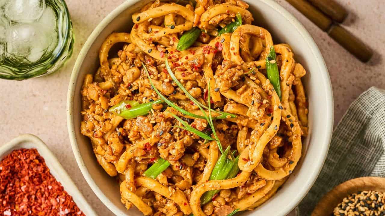 A bowl of stir-fried udon noodles with ground meat, green onions, sesame seeds, and chili flakes, placed on a light surface with chopsticks and seasonings nearby.