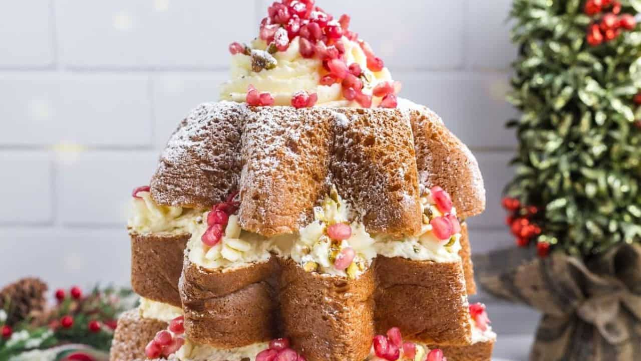 A star-shaped cake layered with cream, topped and filled with pomegranate seeds and powdered sugar, with a small tree decoration in the background.