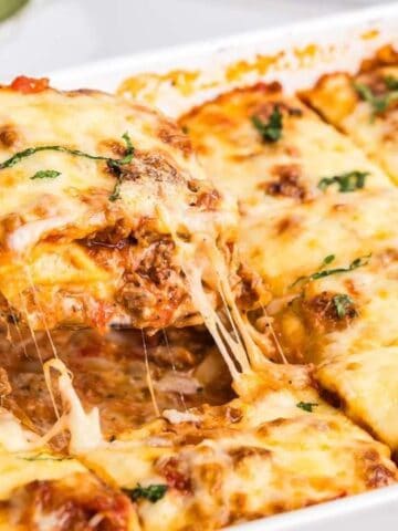A baking dish of cheesy lasagna with one piece being lifted out on a spatula, showing layers of pasta, meat sauce, and melted cheese.