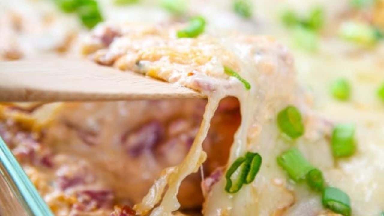 A close-up of a serving spatula lifting a cheesy casserole with chopped green onions from a glass baking dish.