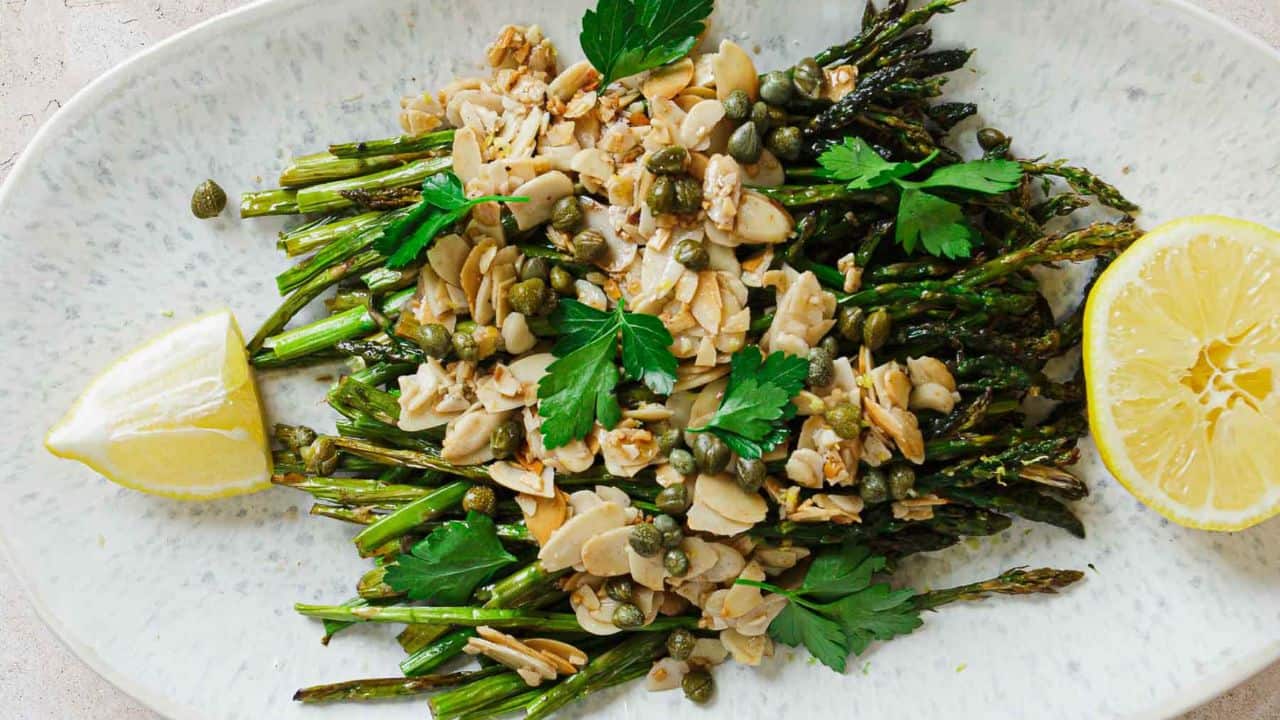 A plate of roasted asparagus topped with almond slices, capers, and parsley. Two lemon wedges are placed on either side of the asparagus. The dish is presented on an oval white plate.
