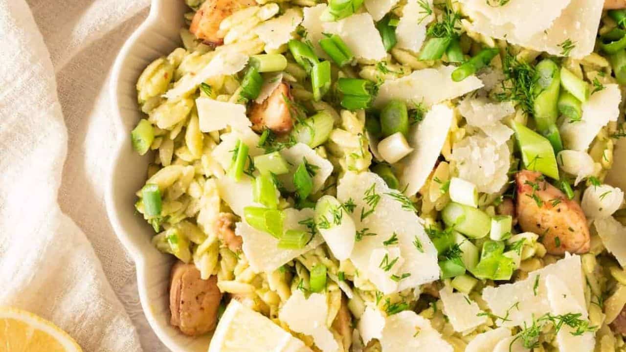 A plate of seafood orzo salad with chunks of squid, shaved parmesan, herbs, served with a fork on a light-colored plate.