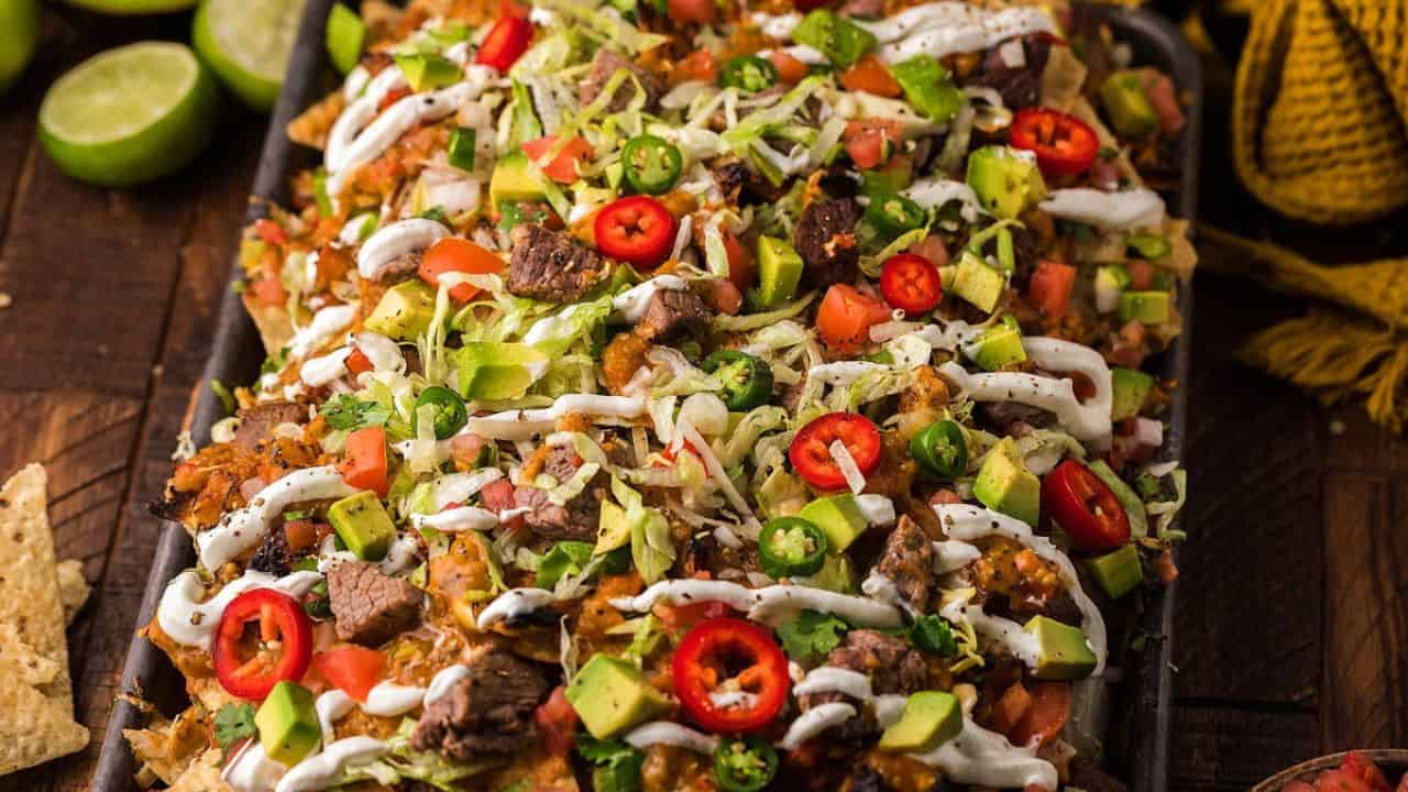 A tray of loaded nachos topped with grilled steak, avocado, tomatoes, jalapeños, shredded lettuce, and drizzled with white sauce. Lime halves and chips are visible in the background.