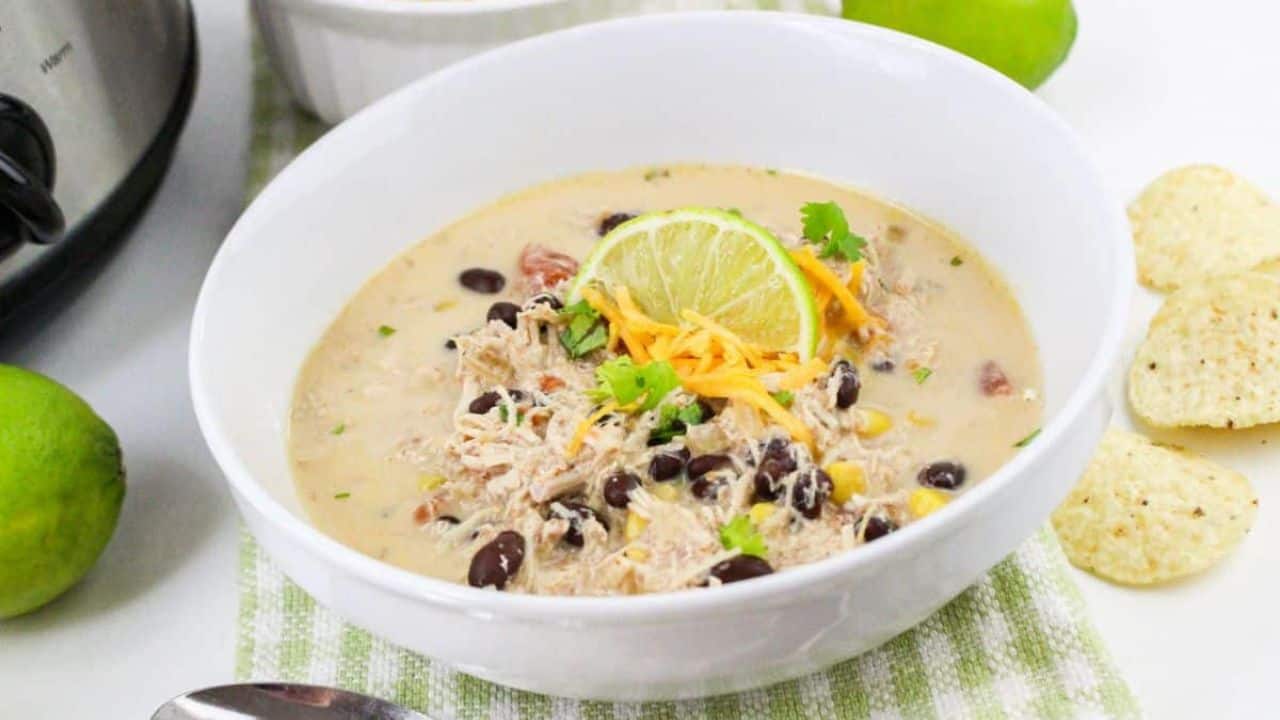 Bowl of creamy chicken soup with shredded chicken, black beans, corn, a lime slice, and cheese on top. Lime, tortilla chips, a spoon, and a slow cooker in the background.