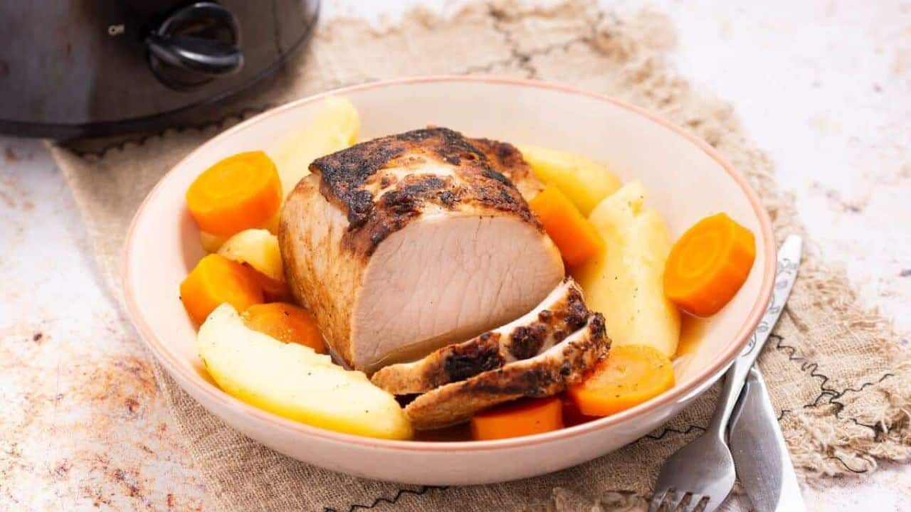 A plate with roasted pork, sliced carrots, and potatoes on a woven mat, with a fork and knife beside the dish. A slow cooker is partially visible in the background.