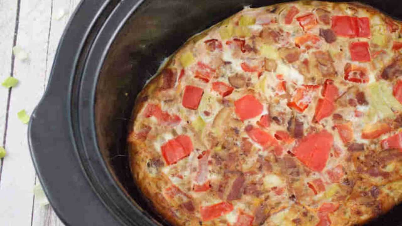 A cooked egg casserole with diced red peppers and other vegetables in a black slow cooker.