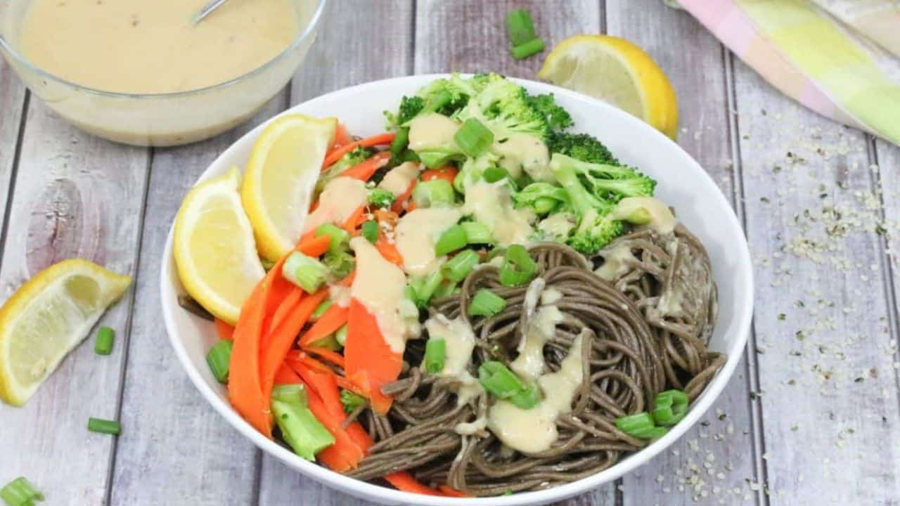 A bowl of soba noodles topped with sliced carrots, broccoli, chopped green onions, lemon wedges, and a drizzle of creamy sauce, with a bowl of extra sauce beside it.