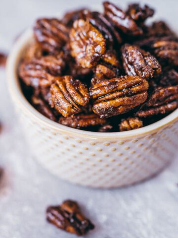A small textured bowl filled with glazed pecans, with a few pecans scattered on a light surface around the bowl.
