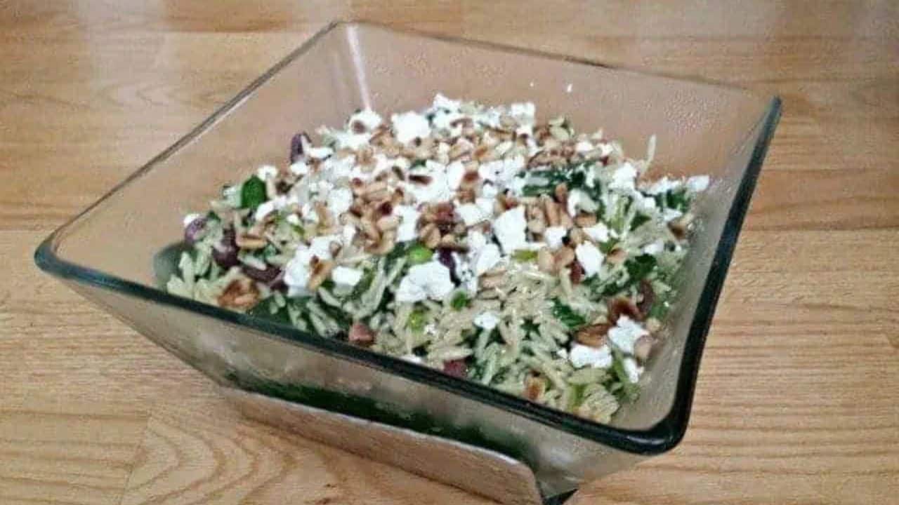 This photo shows a spinach orzo salad in a square glass bowl sitting on a black granite countertop.