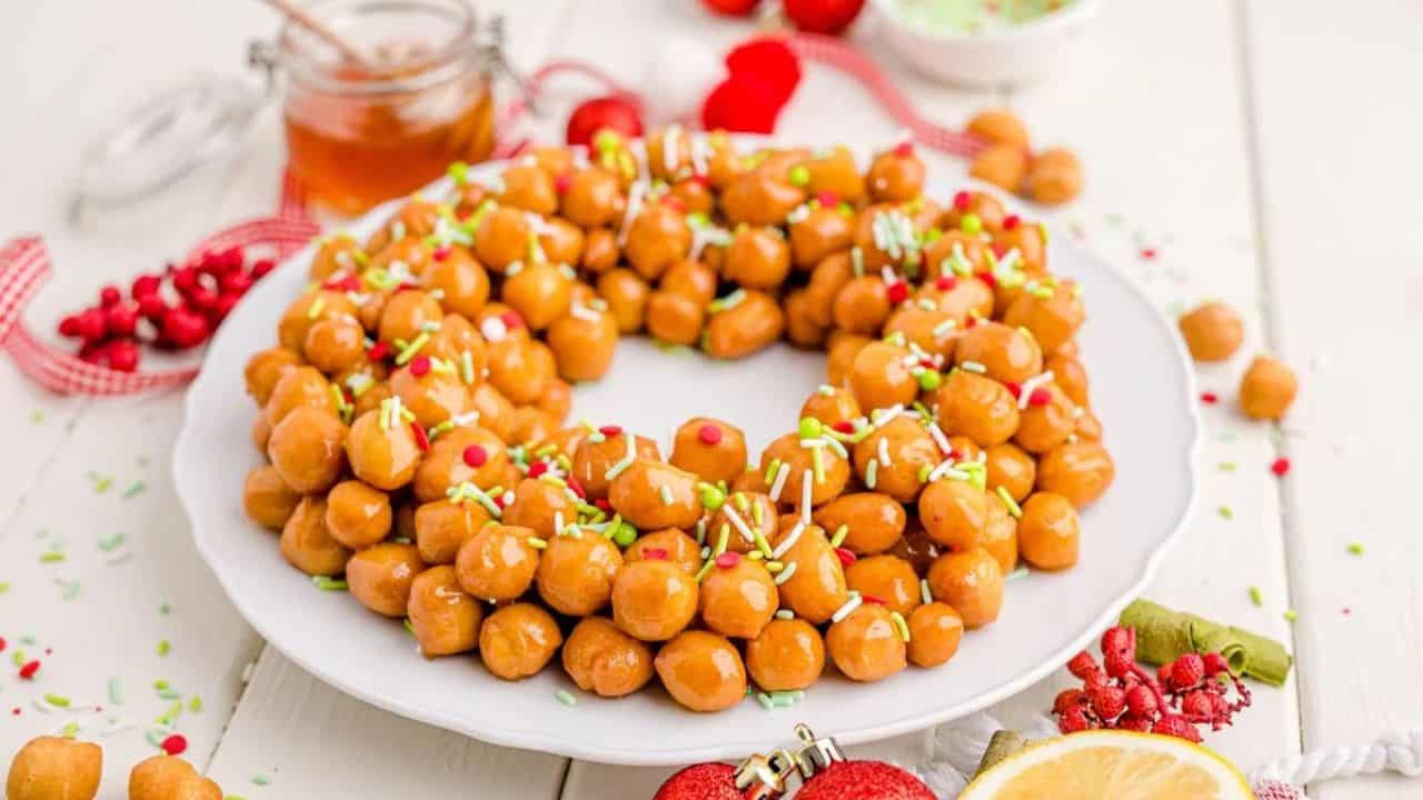 A plate of Italian struffoli arranged in a wreath shape, topped with colorful sprinkles, with honey and lemon nearby on a white table.