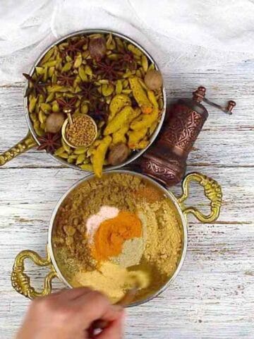 Two brass bowls on a white wooden surface; one contains whole spices, and the other has ground spices being mixed by a hand. A brass pepper grinder is nearby.