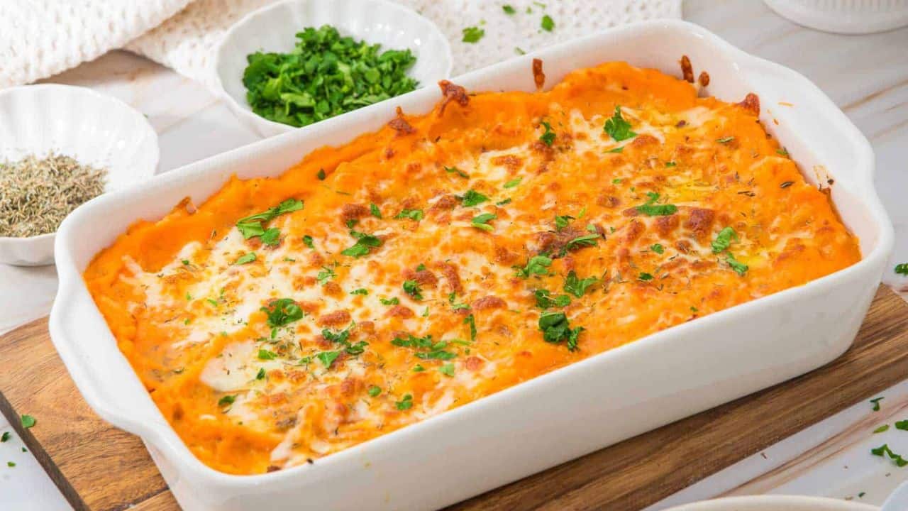 A baked sweet potato casserole topped with melted cheese and chopped parsley, served in a white rectangular dish on a wooden board. Small bowls of fresh herbs and seasonings are visible in the background.