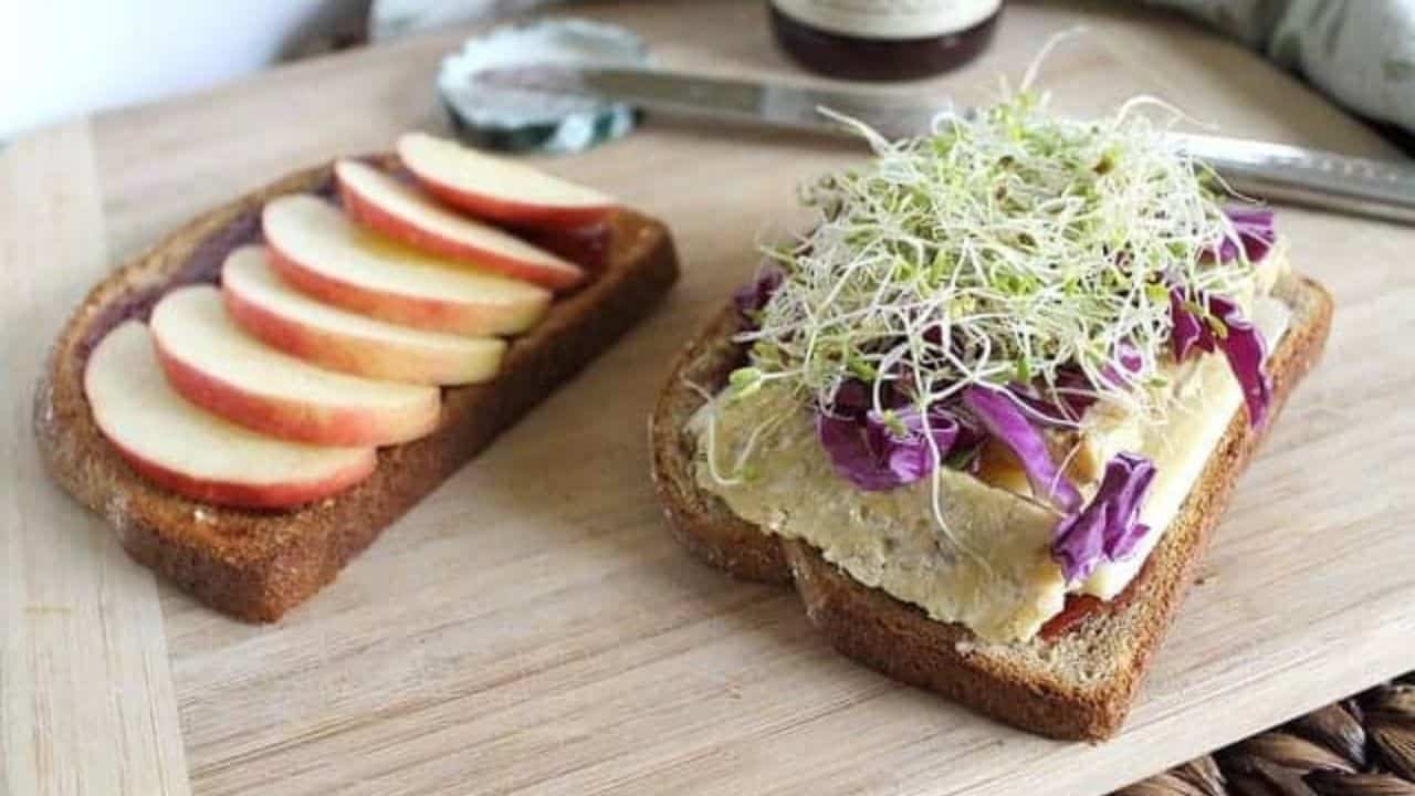 Two slices of wheat bread on a cutting board, one with apple slices, the other with sprouts, purple cabbage, and a thick spread, prepared for a sandwich.