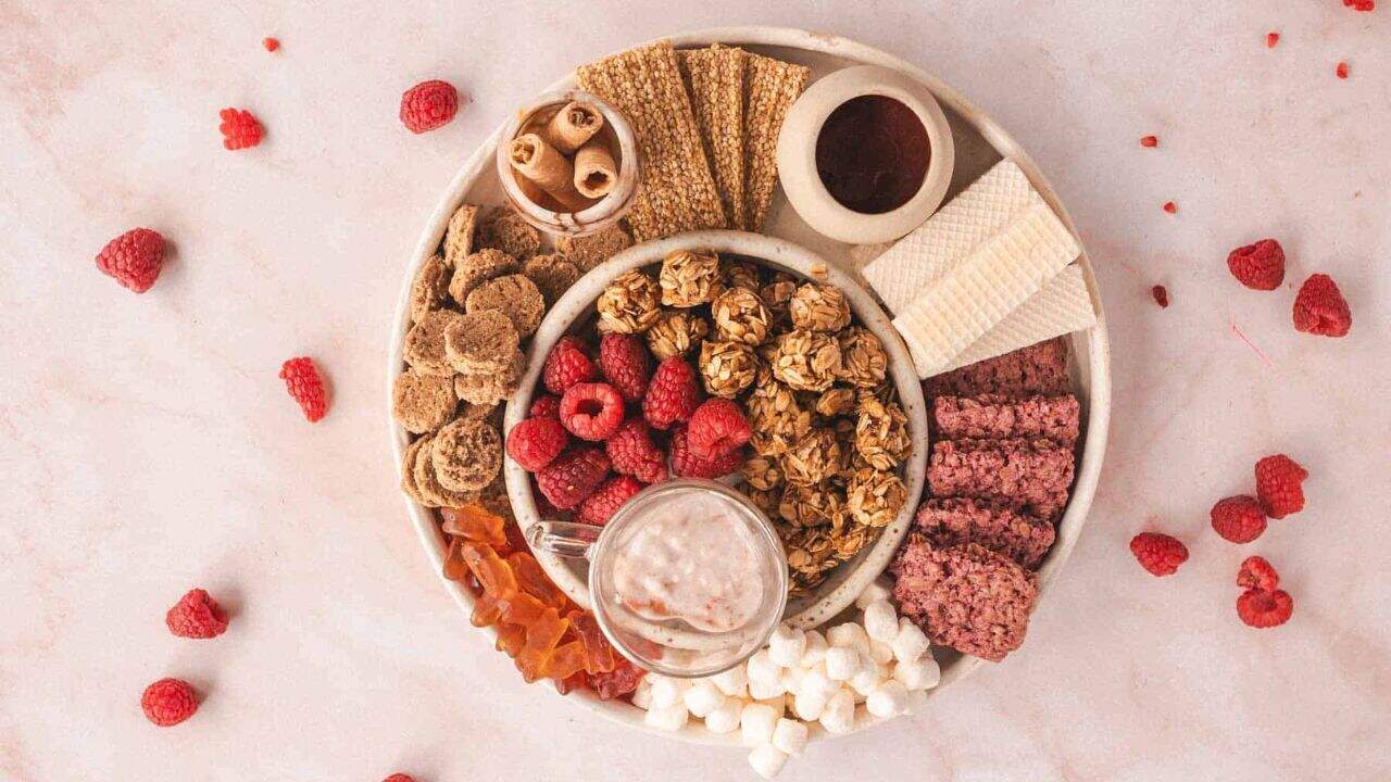 A round platter with raspberries, granola balls, wafers, crispbreads, cookies, marshmallows, and a small mug of smoothie, arranged on a light surface with scattered raspberries.