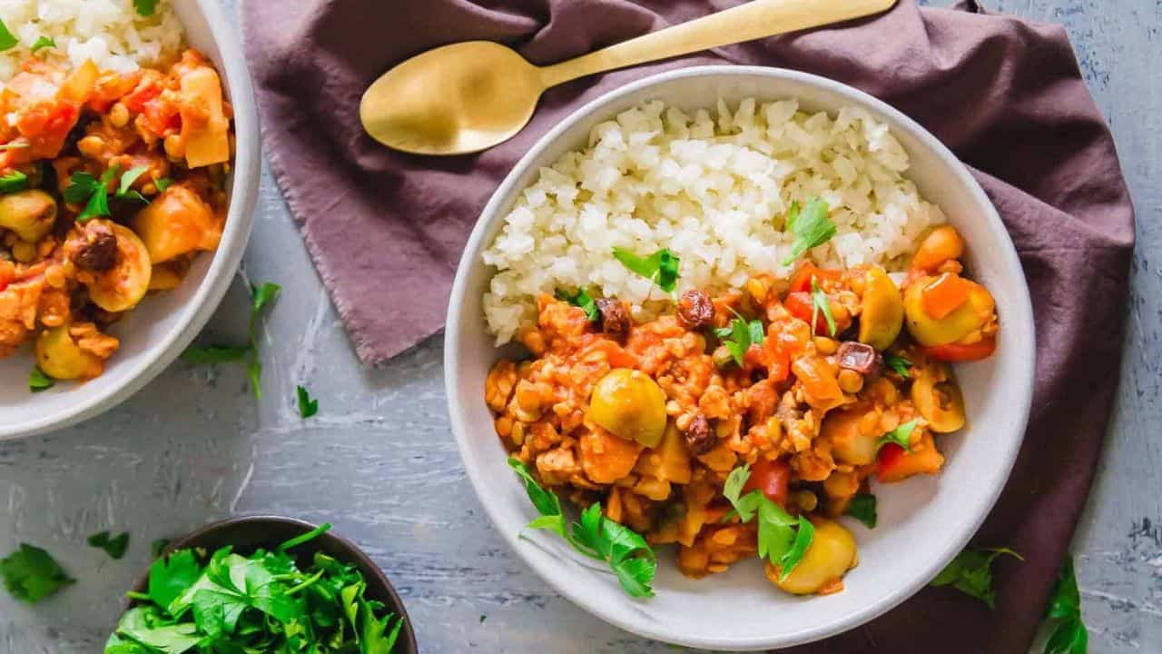 A bowl of white rice served with a vegetable and legume stew, garnished with fresh herbs, placed on a grey surface with a gold spoon and a side dish of greens.