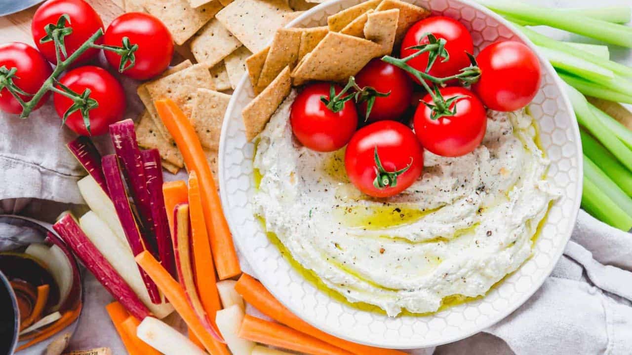 Whipped goat cheese dip topped with olive oil in a bowl with cherry tomatoes and a cheese spreader.