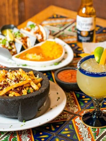 A table with Mexican food including fries in a stone bowl, tacos with rice and beans, tortilla chips, salsa, a margarita, and a bottle of beer on a colorful tiled surface.