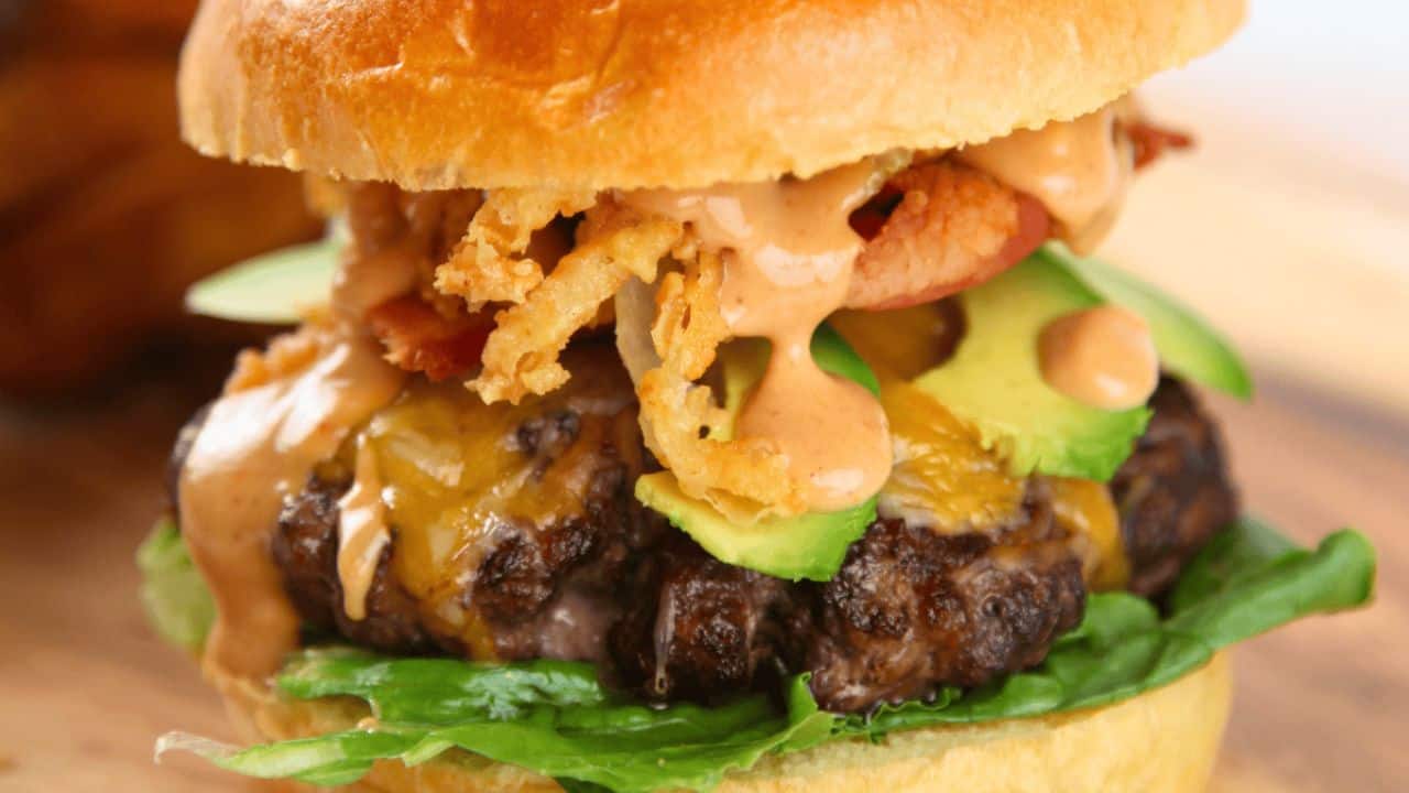 Close-up of a cheeseburger with lettuce, sliced avocado, fried onions, bacon, melted cheese, and sauce in a bun.