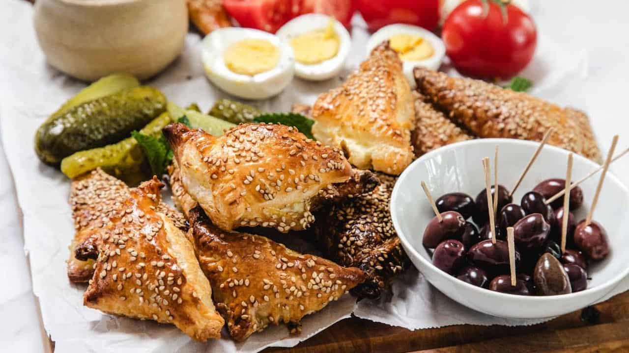 A close-up of golden-brown, triangular puff pastries topped with sesame seeds, stacked on parchment paper, with pickles and olives visible in the background.
