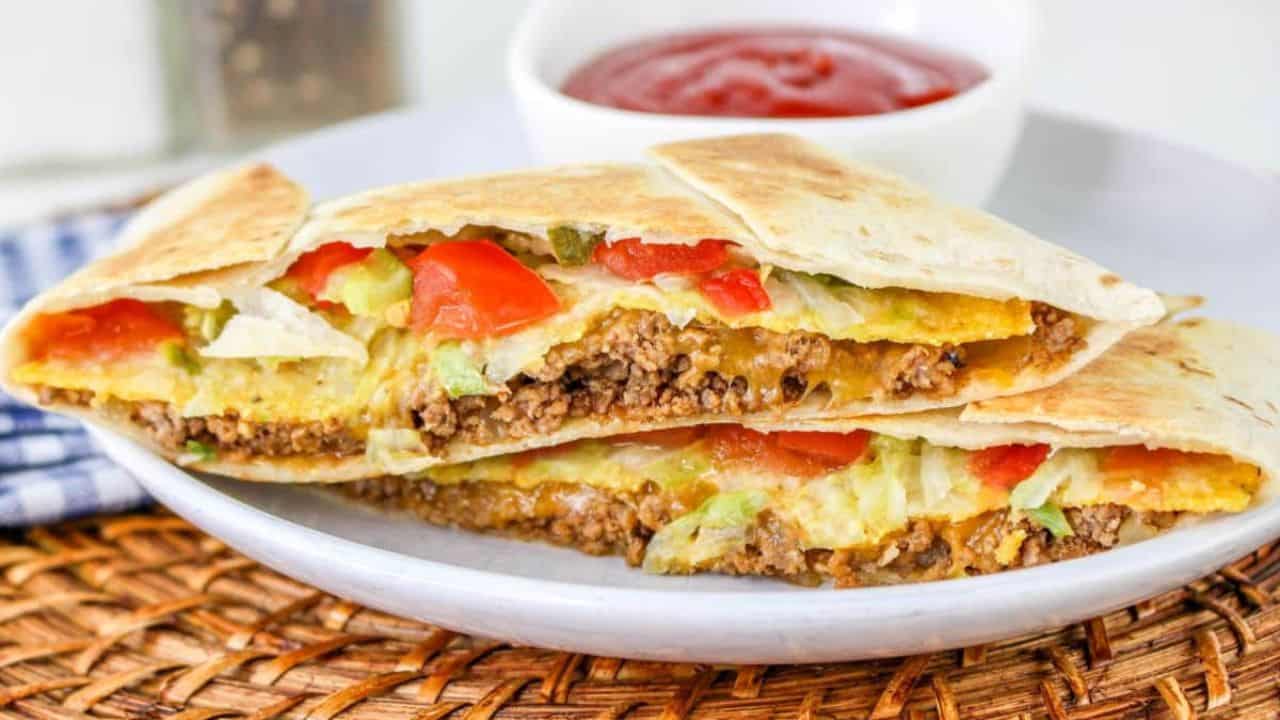 A sliced quesadilla filled with seasoned ground beef, cheese, lettuce, and tomatoes on a plate, accompanied by a small bowl of red sauce.