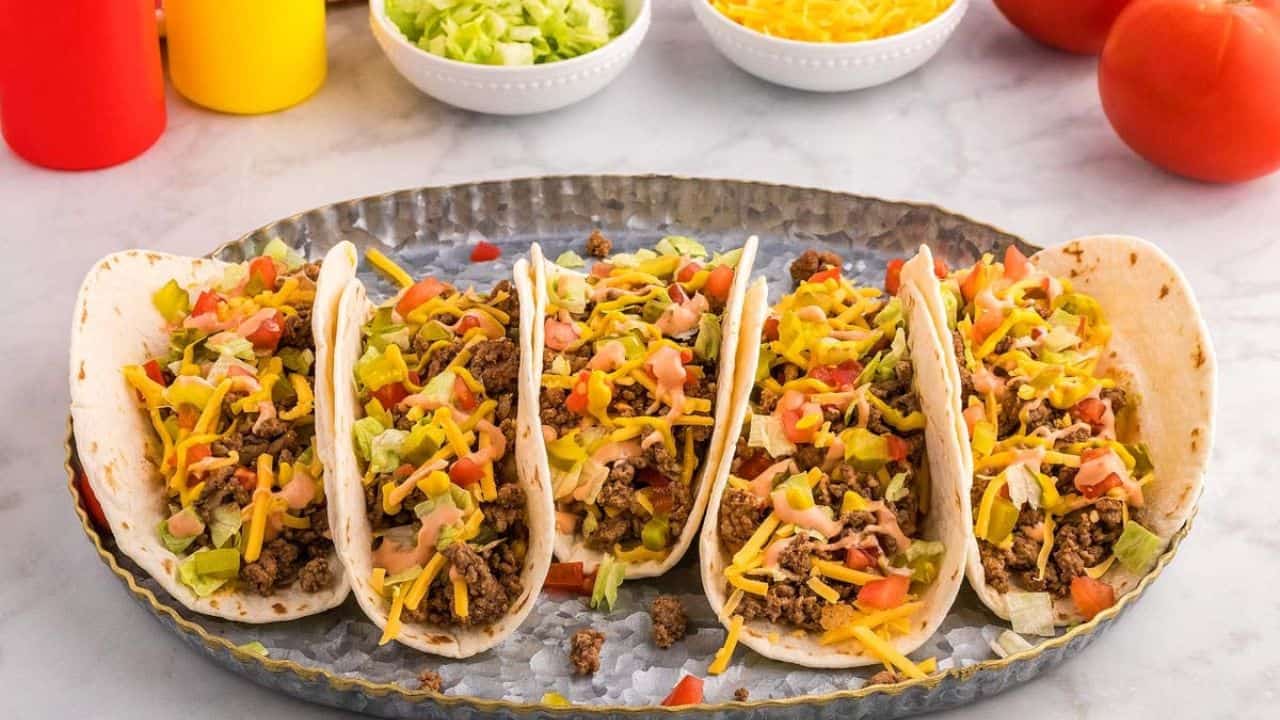 A metal tray holding five beef tacos topped with lettuce, tomato, shredded cheese, and sauce. Bowls of lettuce and cheese, tomatoes, and condiment bottles are in the background.