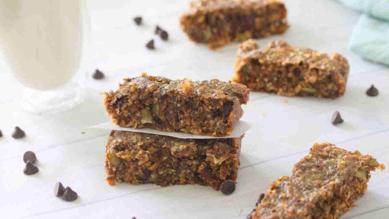 Two granola bars are stacked on parchment paper, with more bars and scattered chocolate chips on a white surface. A glass of milk is in the background.