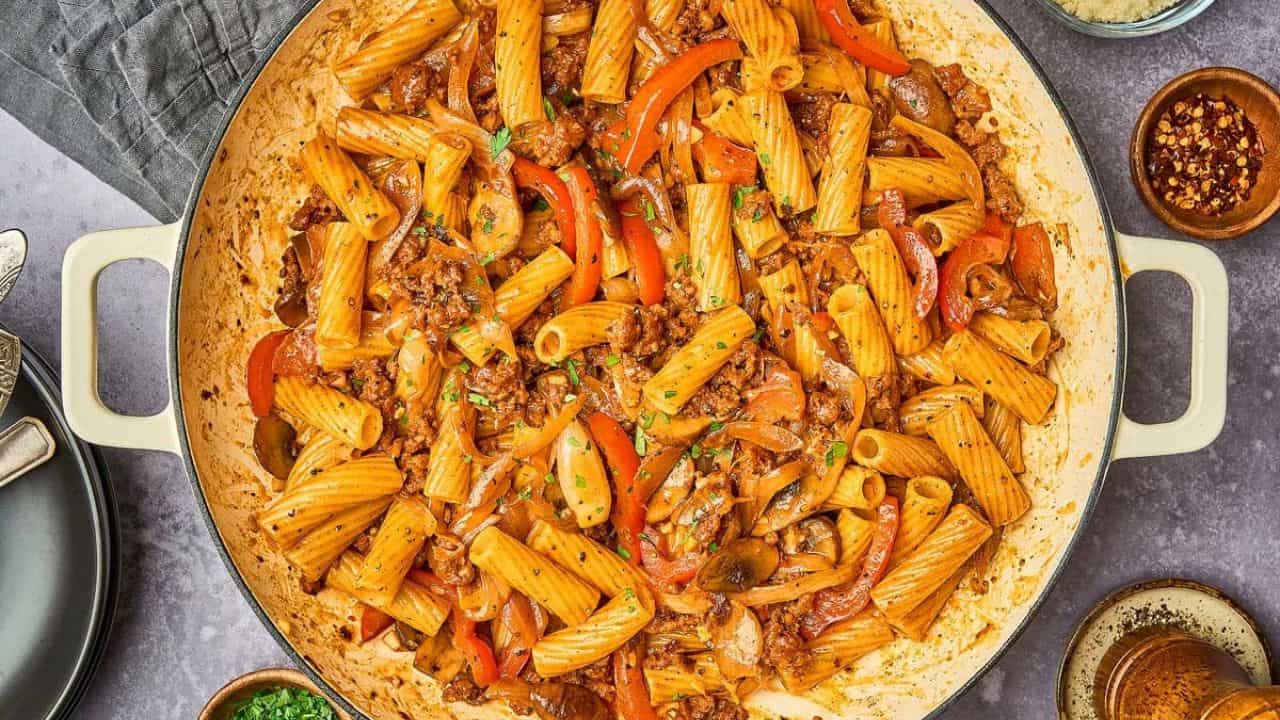 Large skillet filled with rigatoni pasta, ground meat, red bell peppers, and a red tomato-based sauce, surrounded by small bowls of spices and grated cheese.