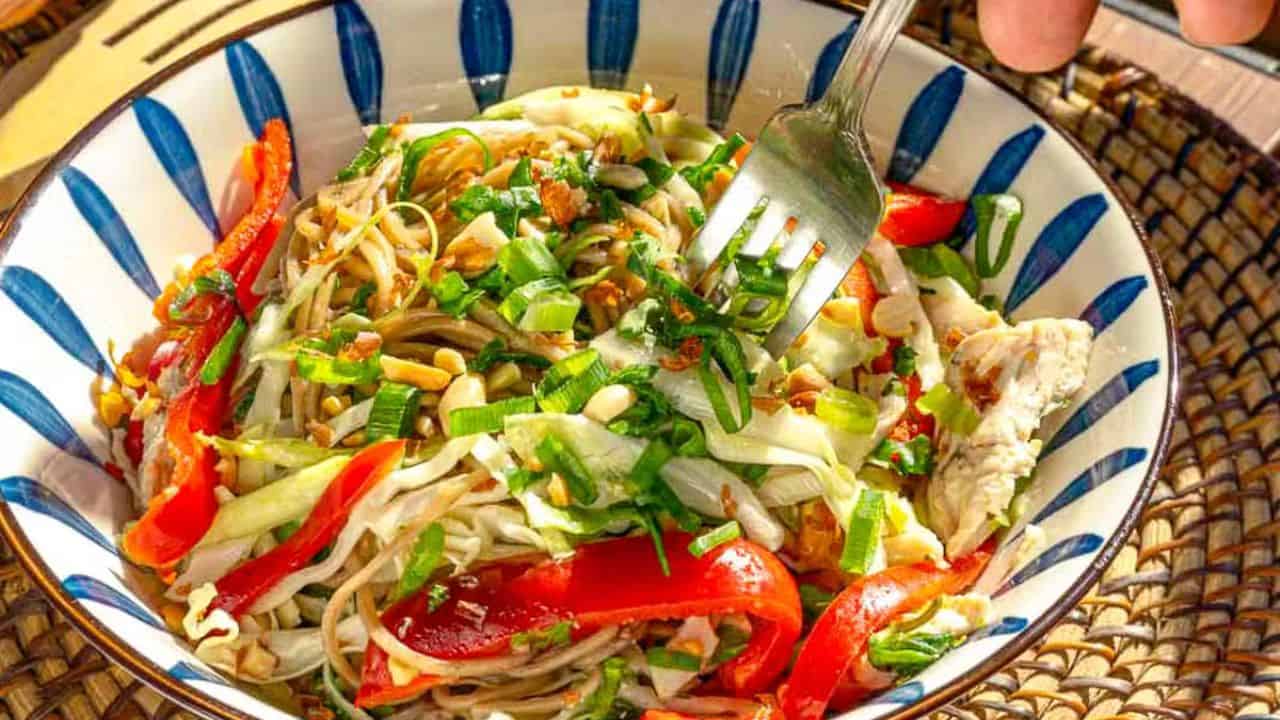 A colorful salad in a striped bowl, featuring mixed vegetables such as red bell peppers and shredded cabbage, garnished with crushed nuts and fresh herbs. A fork is seen lifting a portion, emphasizing the dish's vibrant, fresh ingredients.