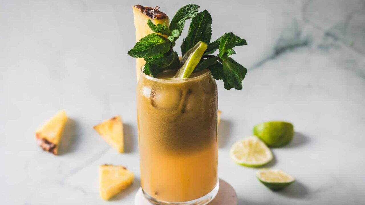A tall glass of iced brownish-orange beverage garnished with a sprig of fresh mint, a pineapple wedge, and a lime wedge. Slices of pineapple and lime are scattered on a marble surface in the background.