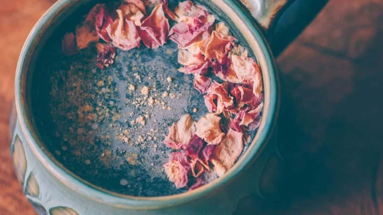 A ceramic mug filled with a dark beverage, topped with pink dried flower petals and a sprinkle of light-colored powder.