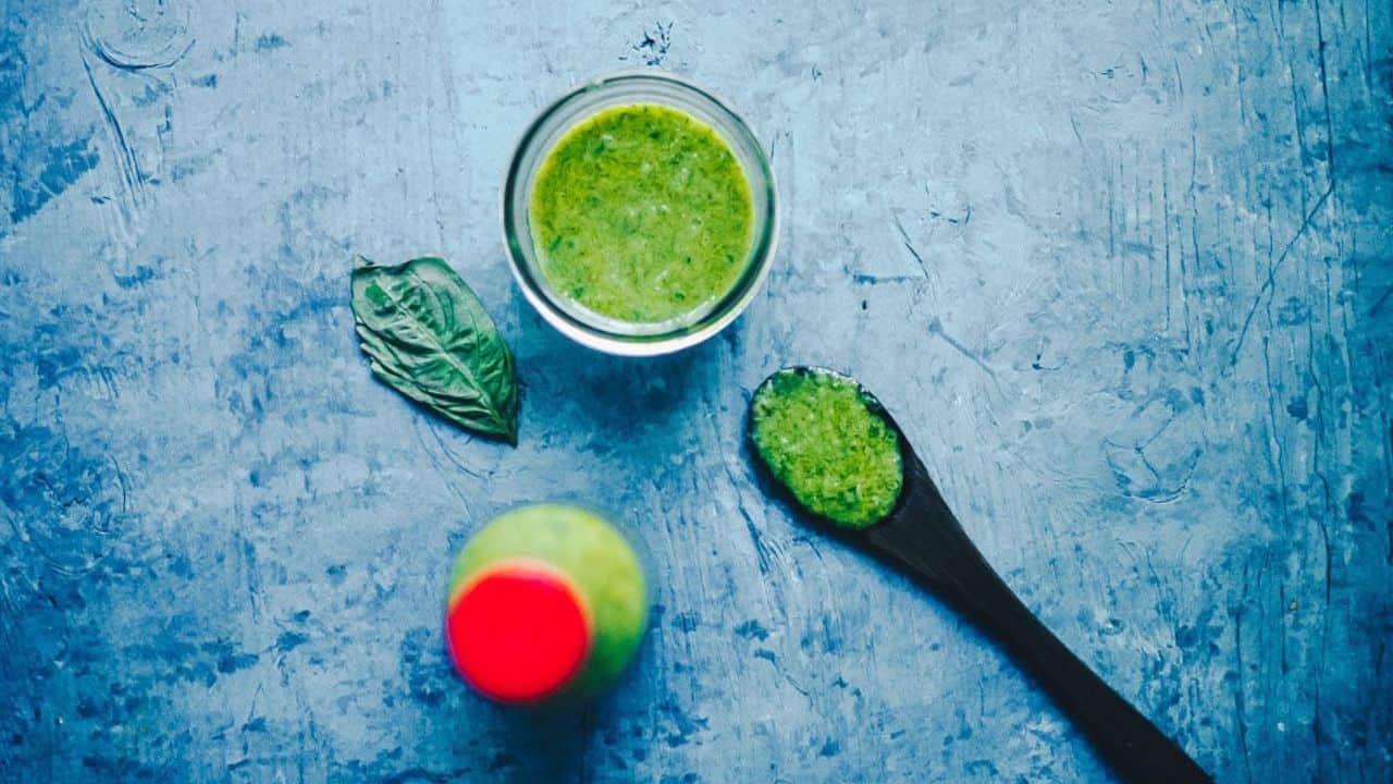 A glass of green smoothie, a leafy herb, and a spoonful of the smoothie are arranged on a textured blue surface next to a bottle with a red cap.