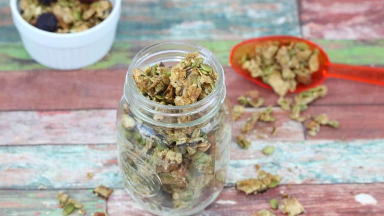 A glass jar filled with granola sits on a rustic wooden surface, with a red spoon and a white ramekin of granola in the background.