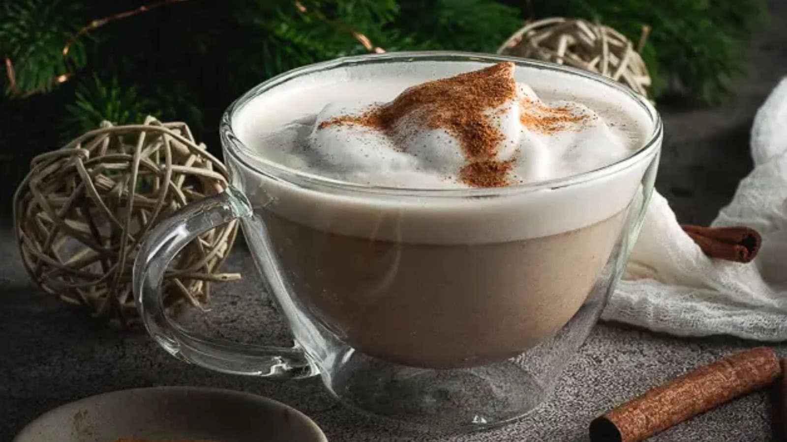 A clear glass mug filled with a frothy coffee beverage topped with foam and sprinkled cinnamon, with decorative items and a cinnamon stick nearby.