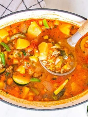 A pot of vegetable soup with chunks of zucchini, potatoes, carrots, green beans, and ground meat, with a ladle inside.