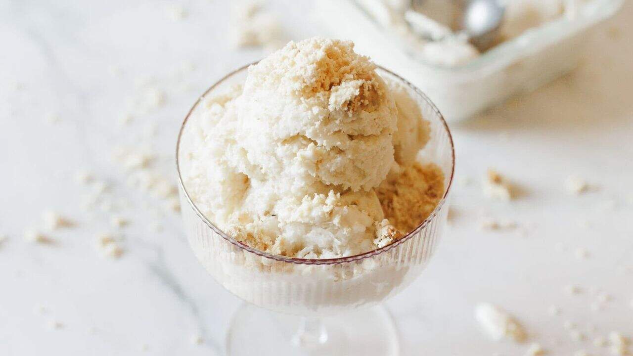 A glass dessert dish filled with scoops of vanilla ice cream, topped with crumbled cookies, sits on a white surface with crumbs scattered around.