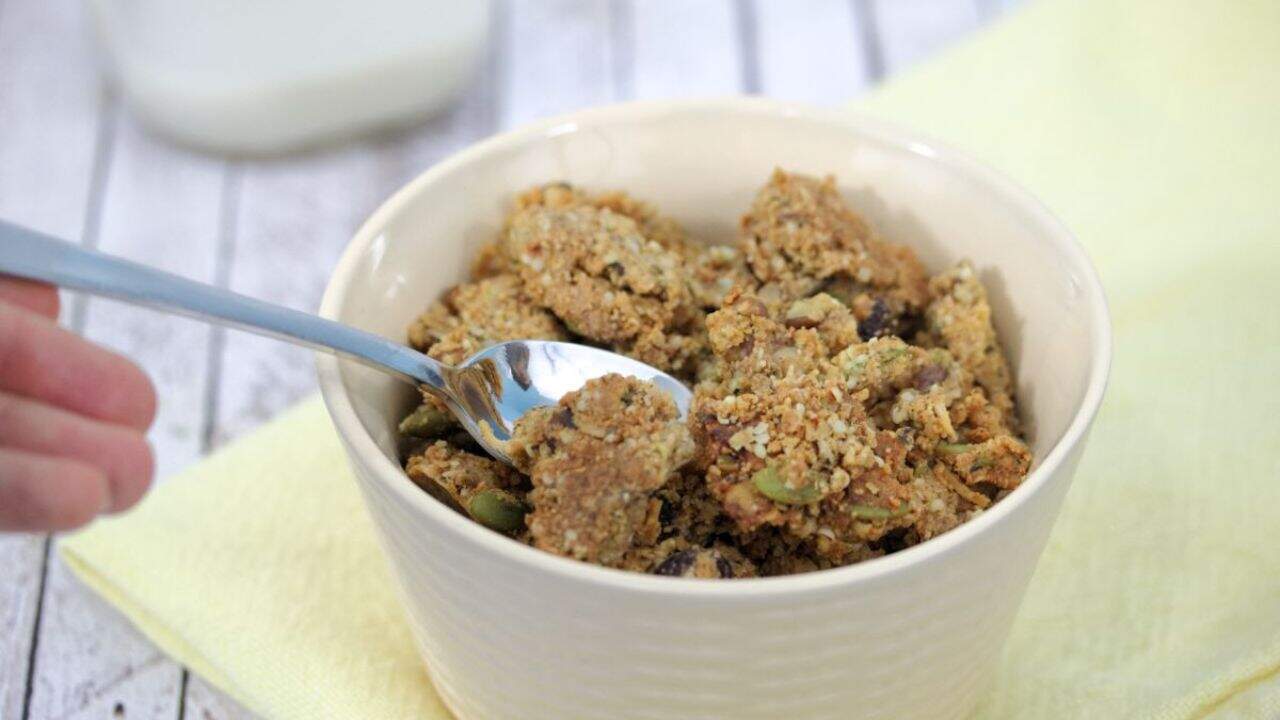 A bowl of granola clusters with seeds and nuts, with a spoon scooping some up. A glass of milk is in the background on a yellow napkin.