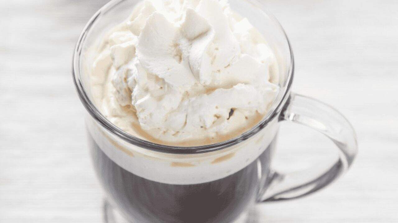 A clear glass mug filled with coffee topped with a generous swirl of whipped cream, placed on a light surface.