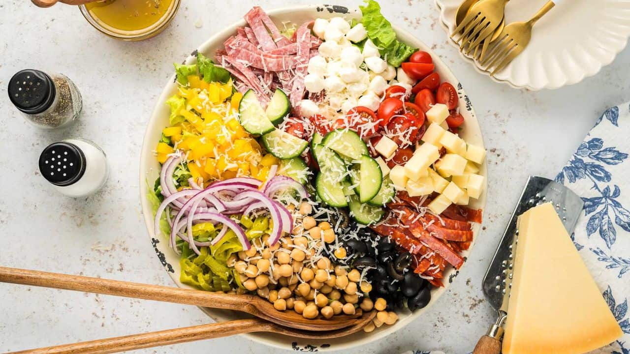 A large bowl of salad with sliced cucumbers, tomatoes, red onions, bell peppers, chickpeas, olives, cheese cubes, salami, and lettuce, with wooden salad servers and seasonings nearby.
