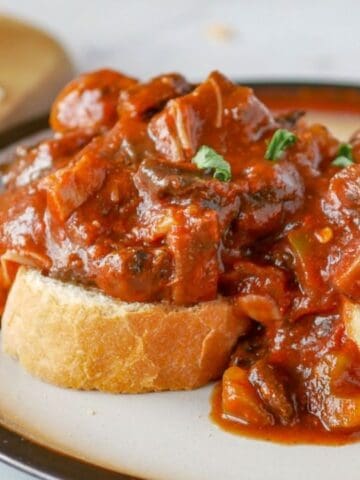 Two slices of bread topped with a chunky tomato-based stew are served on a plate, with fresh greens and a cutting board in the background.