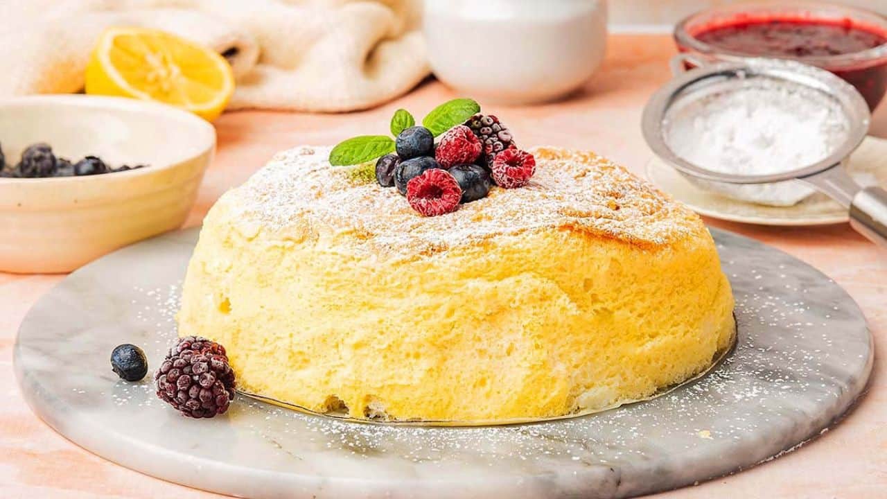 A fluffy Japanese soufflé pancake topped with powdered sugar, raspberries, blueberries, and a mint sprig, displayed on a marble platter with baking items in the background.