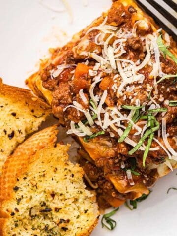 A plate with a serving of lasagna topped with shredded cheese and herbs, two slices of garlic bread, and a fork on the side.