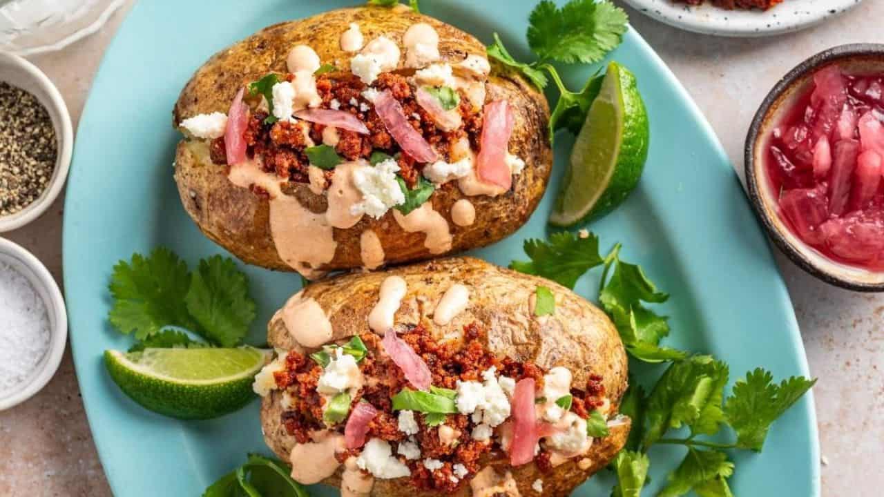 Two baked potatoes topped with crumbled chorizo, cheese, pickled onions, and creamy sauce are served on a blue plate with lime wedges and cilantro.
