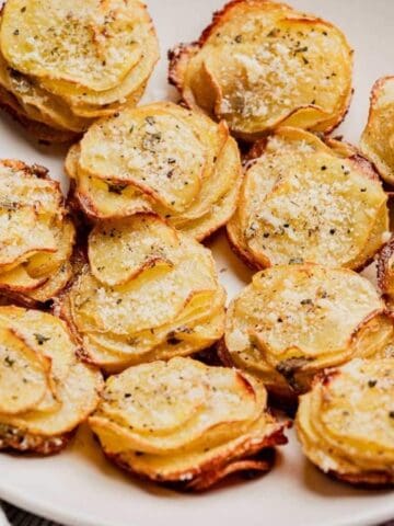 A white plate holds several stacks of thinly sliced, baked potatoes topped with herbs and grated cheese.