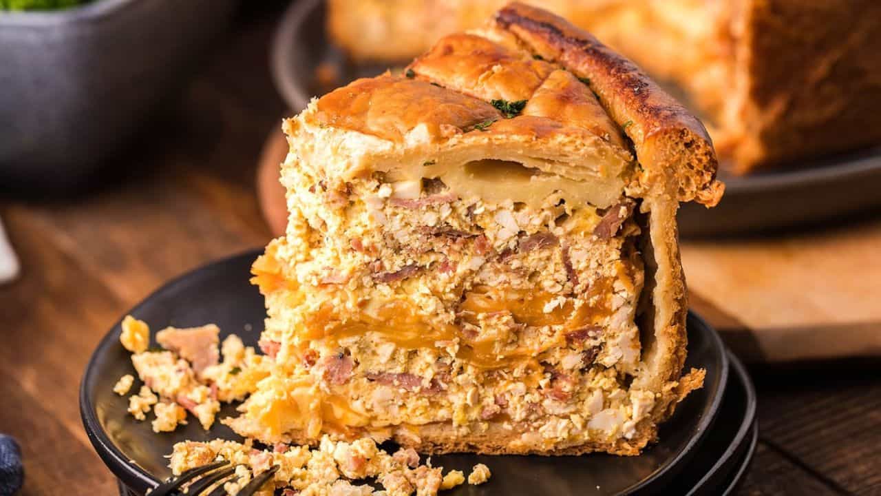 A thick slice of savory pie with a golden crust and visible layers of meat, cheese, and eggs sits on a black plate with a fork.
