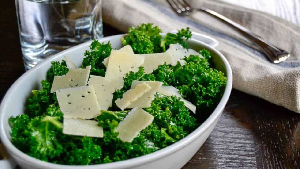 A white bowl filled with fresh kale topped with shaved cheese slices creates a delightful salad perfect for impressing guests. A glass of water, fork, and a napkin are elegantly placed on the dark wooden table.
