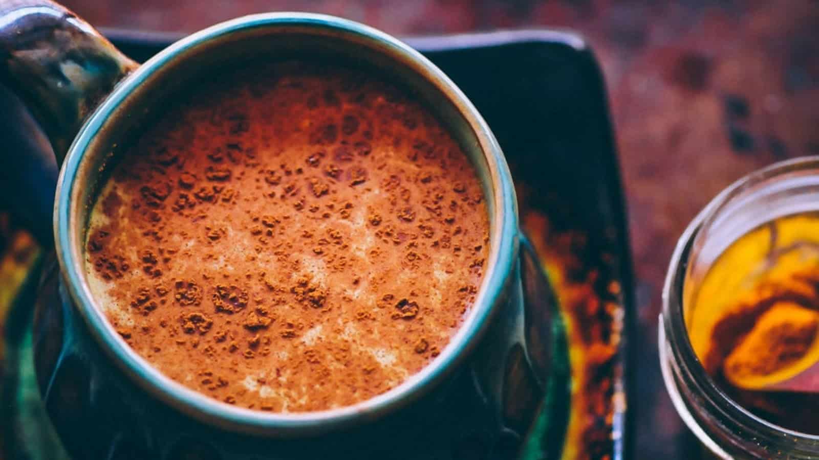 A close-up of a ceramic mug filled with a frothy, brown liquid, possibly coffee or hot chocolate, next to a glass jar on a dark surface.