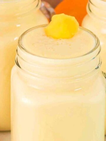Three glass mugs filled with a creamy yellow smoothie, with a dollop of fruit on top of the front mug, and a banana and oranges nearby.