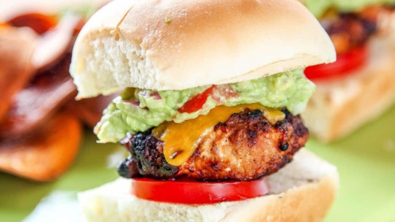 A grilled chicken burger with cheddar cheese, tomato slices, and guacamole on a white bun.